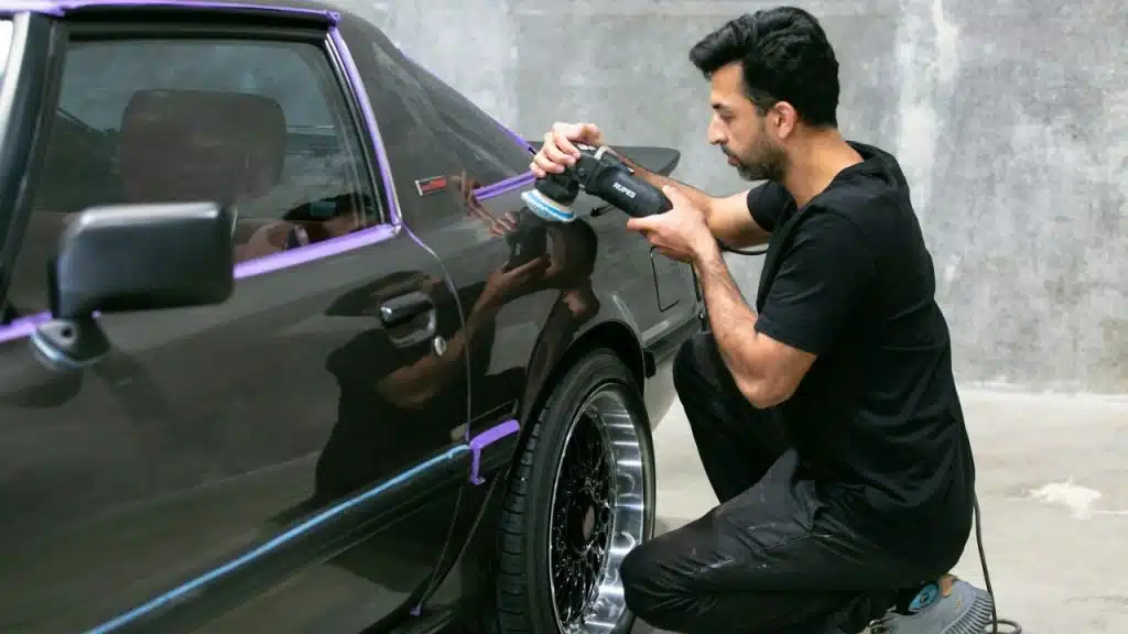 A man kneels beside a car and uses a buffer to detail it.