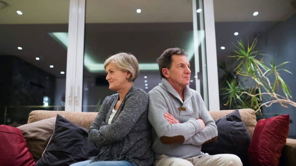 An older man and woman sit back to back on a couch with their arms crossed.