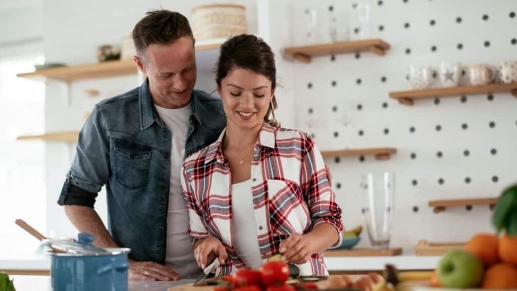 A happy couple stands close together in a kitchen. The woman is chopping vegetables, and the man is watching her.