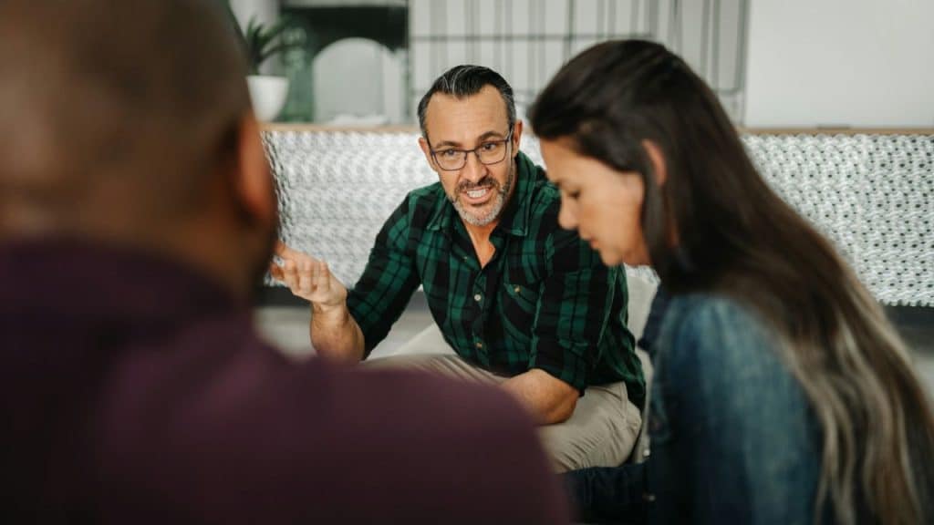 A man in a flannel shirt and glasses talks and gestures, while a woman and another person listen.