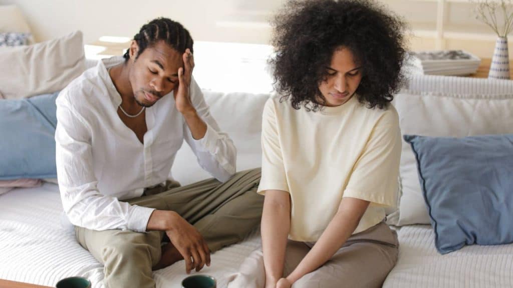 A man and a woman sit on a sofa, both looking down and away from each other. The man is holding his head in his hand.