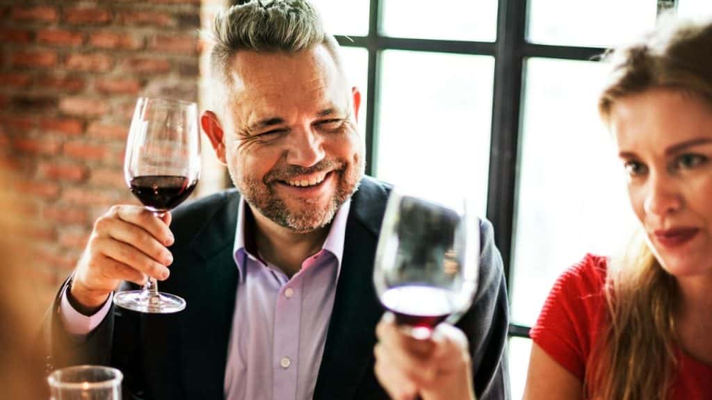 A man with a salt-and-pepper beard is smiling and holding a glass of red wine.