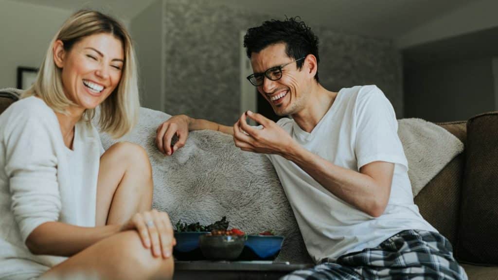 Blonde woman and man in glasses laughing while sitting on a couch with bowls of food.
