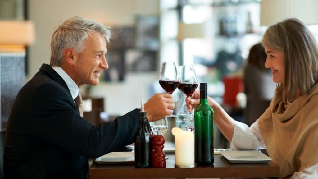 A smiling older couple is clinking wine glasses across a table in a restaurant.