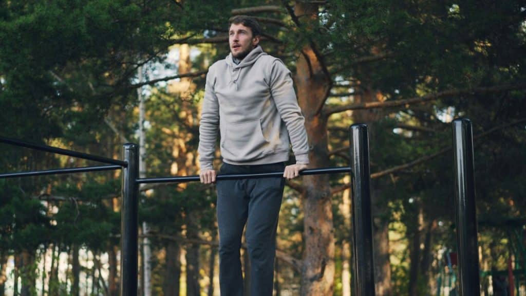 A man in a hoodie is doing a dip exercise on black parallel bars outdoors.