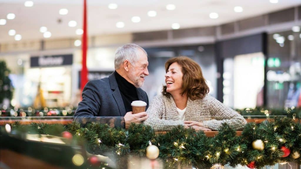 A laughing, older couple is enjoying a coffee break in a shopping mall decorated with festive garlands.