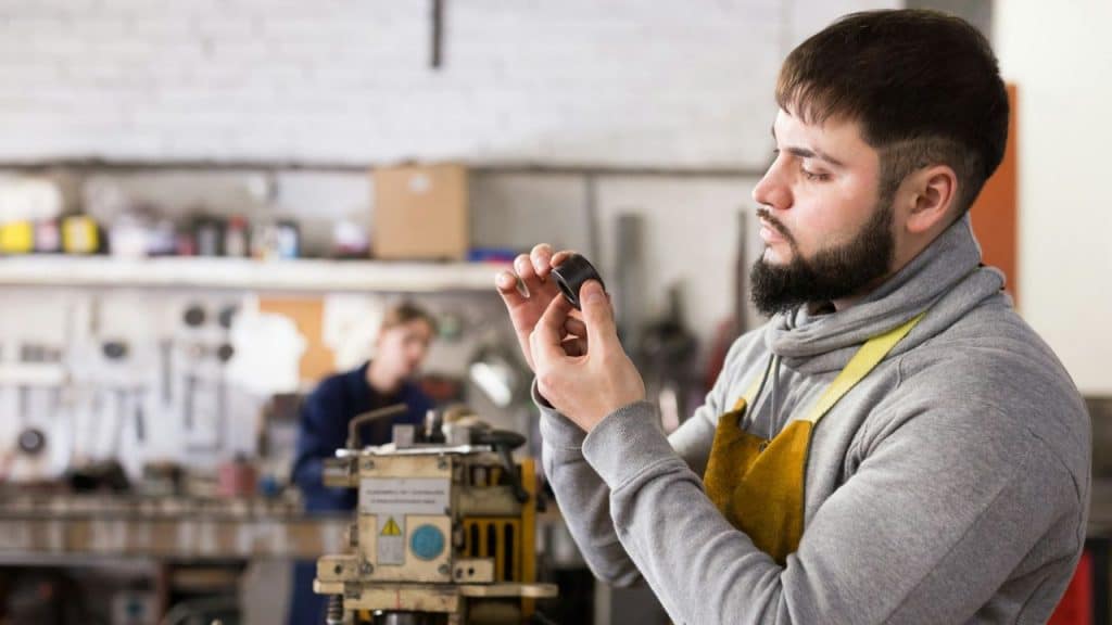 A man with a beard inspects a small part in a busy workshop.