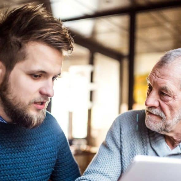 A young, bearded man and an older man are looking at a tablet together and talking indoors.