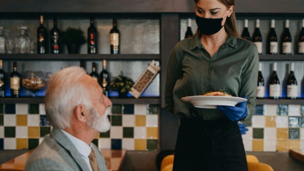 A waitress wearing a black mask and blue gloves serves a plate to an older man in a restaurant.