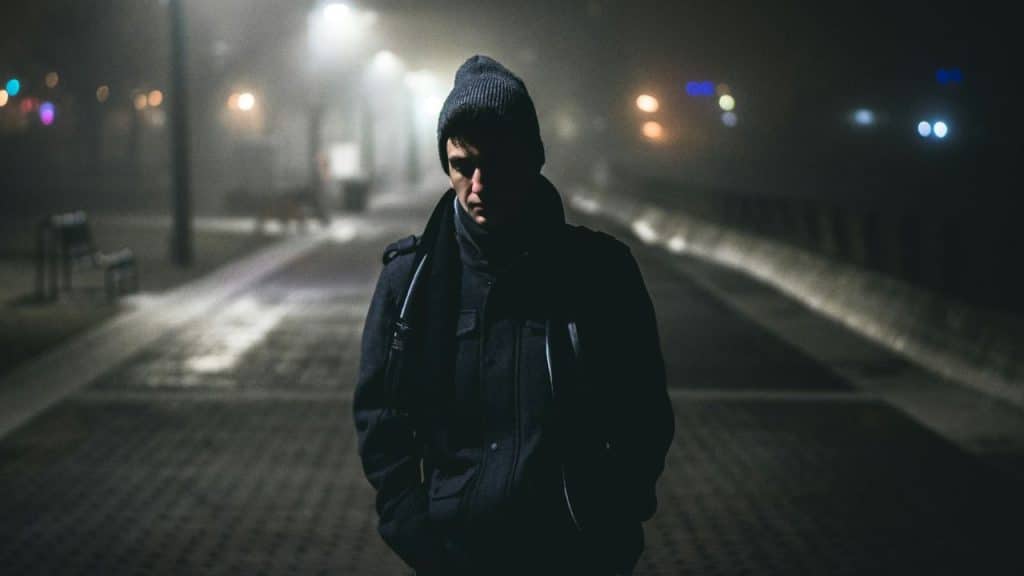 A lonely man in a coat and beanie walks on a dark, foggy street.