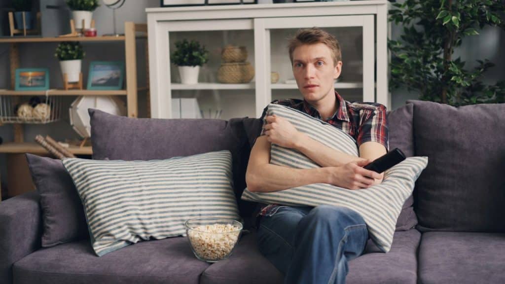 A sad-looking man sits on a couch, holding a pillow, with a bowl of popcorn and a remote control.