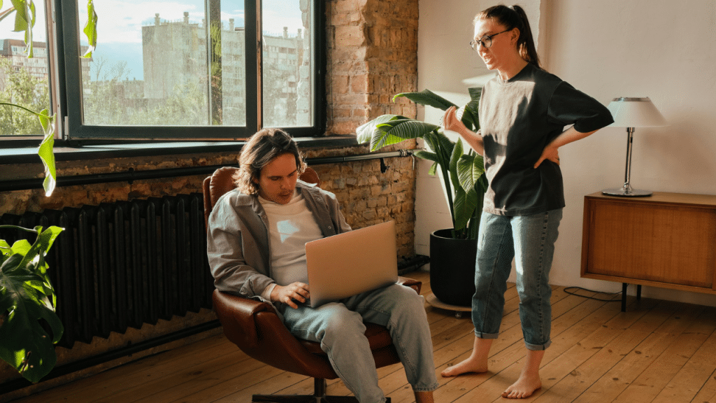 A woman with her hand on her hip stands next to a man sitting in a chair with his laptop.