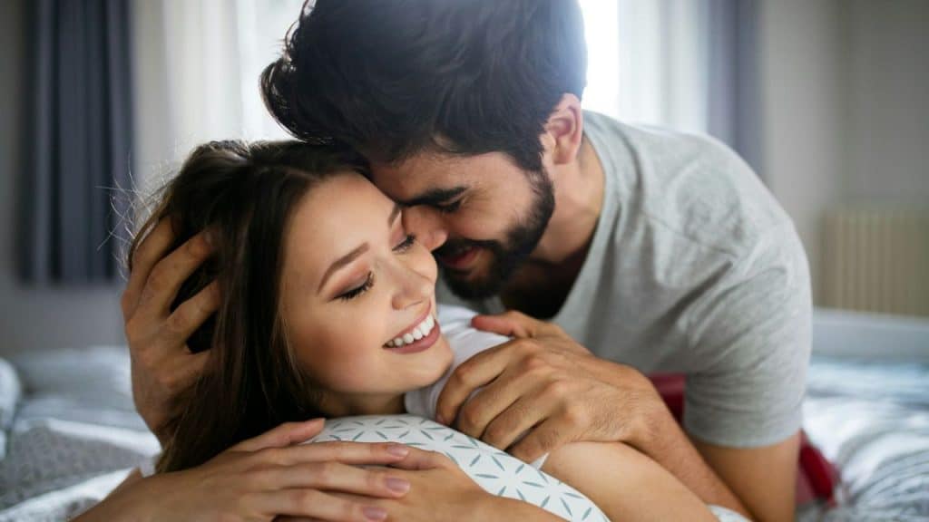 A smiling couple is lying in bed, hugging each other and looking at each other.