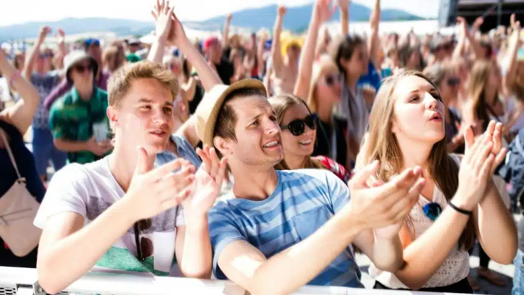 A large crowd of young people cheering and clapping at an outdoor music festival.
