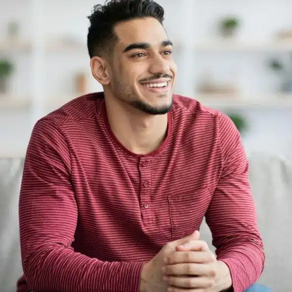 A handsome, young Middle Eastern man with a beard sits on a couch, smiling and looking off to the side.