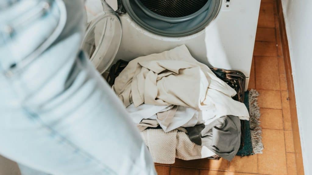 A person stands near an open washing machine, which has a basket full of clothes next to it.