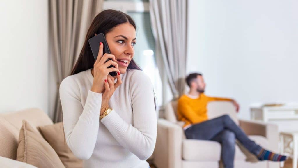 A woman on the phone smiles and looks away from a man sitting on a chair in the background.