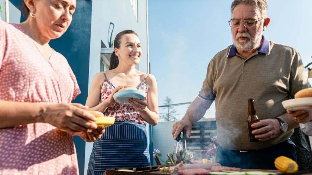 A senior man grills meat on a barbecue while two women look on.