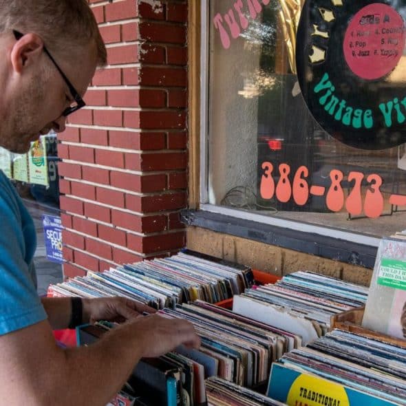 A man in a blue shirt is browsing through a box of vinyl records outside a vintage store.