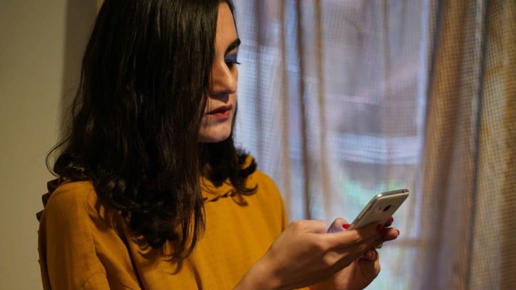A woman with dark hair and a yellow shirt looks at her phone with a focused expression.