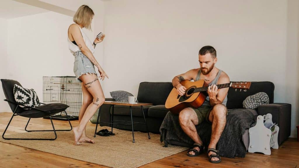 A woman on her phone and a man playing guitar in a living room.