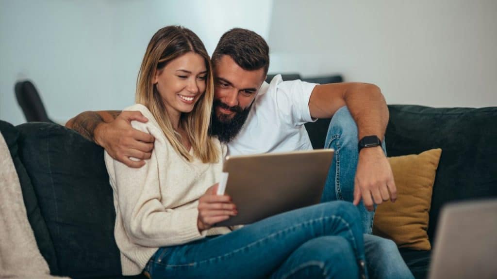 A couple sits close on a couch, smiling as they look at a tablet together.