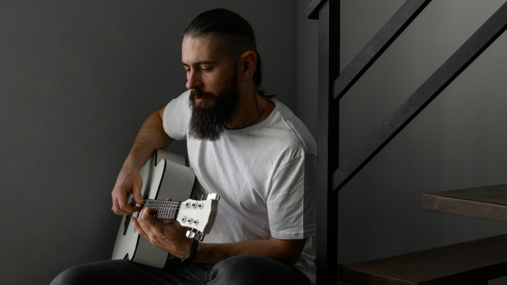 A sad man with a beard and ponytail sits and plays an acoustic guitar.