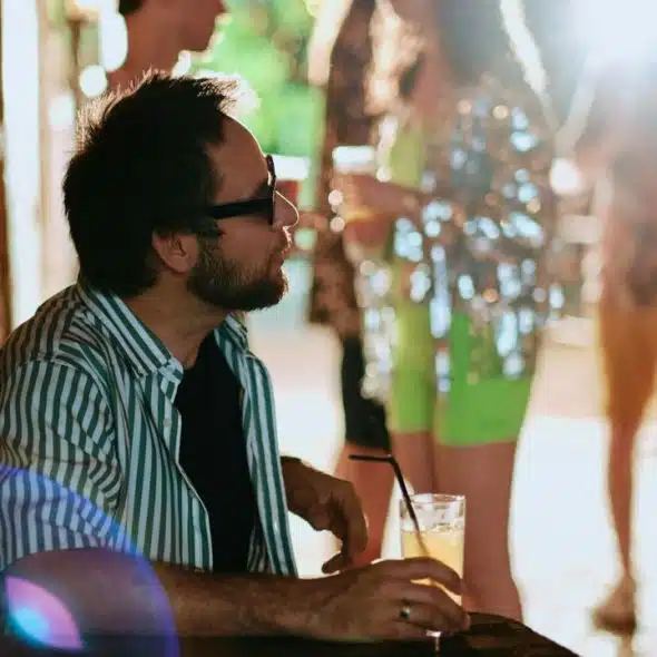 A man sits alone with a drink at a party, looking to the side.