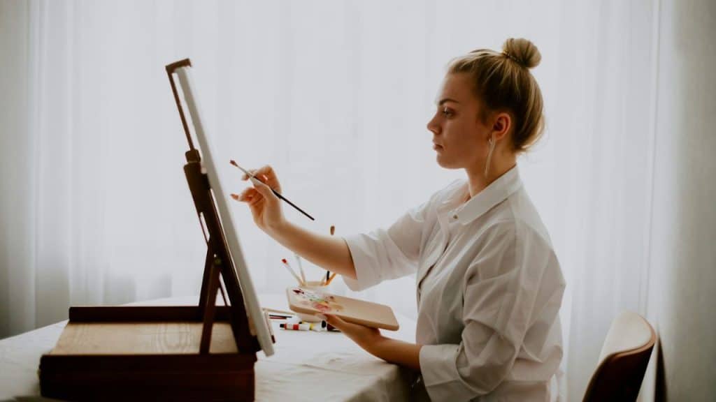 A young woman in a white shirt paints on a canvas while holding a palette.