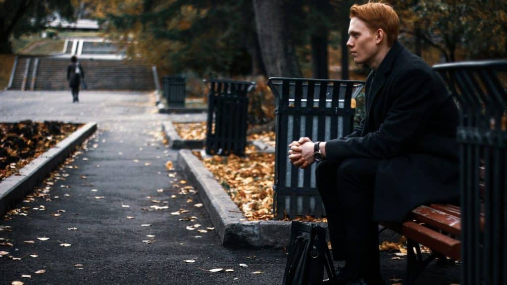 A serious-looking man with red hair sits on a park bench, gazing into the distance.