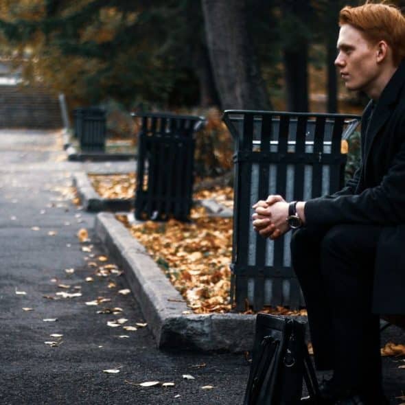 A serious-looking man with red hair sits on a park bench, gazing into the distance.