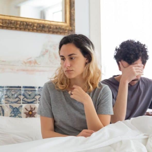 A man and a woman sit in bed. The man covers his face with his hand.