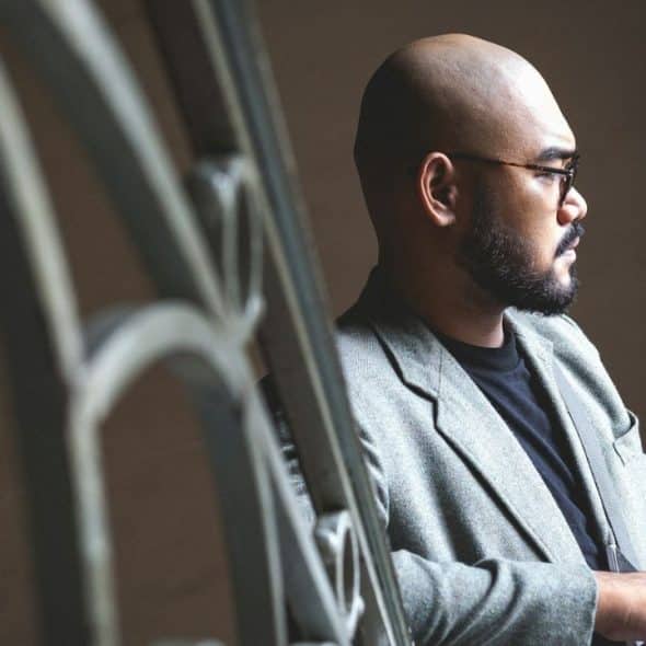 A bald man with a beard and glasses looks to the side while leaning on a metal railing.