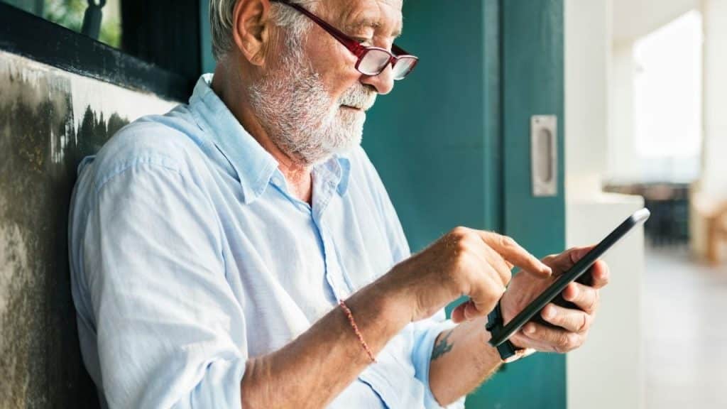 A gray-bearded older man wearing glasses uses a phone outdoors.