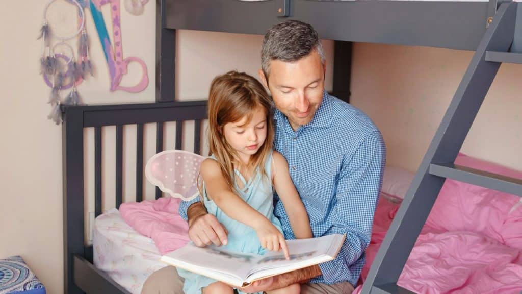 A father and daughter, who is wearing fairy wings, sit on a bed reading a book.