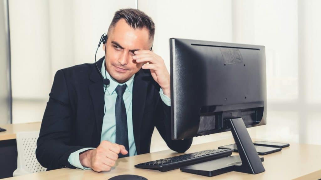 A stressed-looking man in a suit and headset sits at a desk, rubbing his eye.