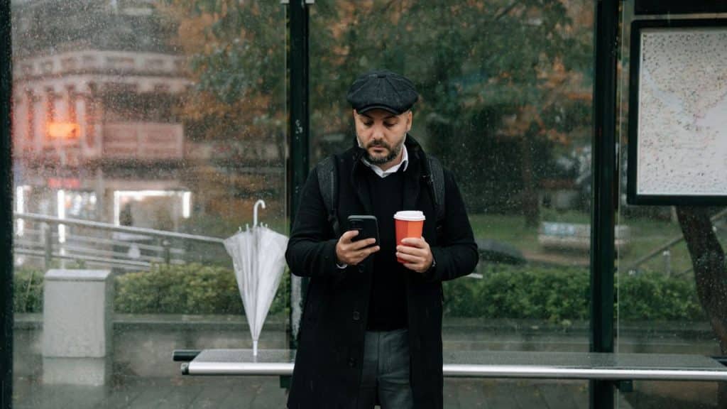 A man in a black coat and cap checks his phone at a bus stop on a rainy day.