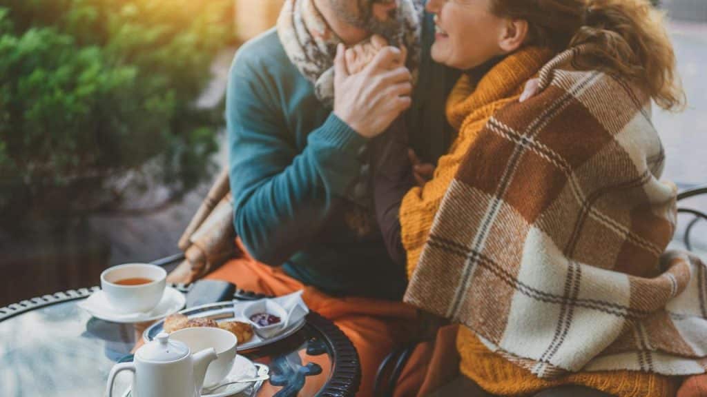 A couple sitting close together under a blanket, holding hands at an outdoor café table with tea and pastries.