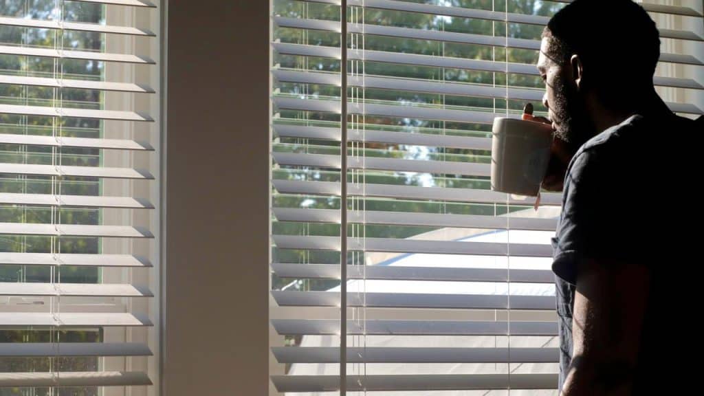 A man drinking from a mug while looking out the window through blinds.