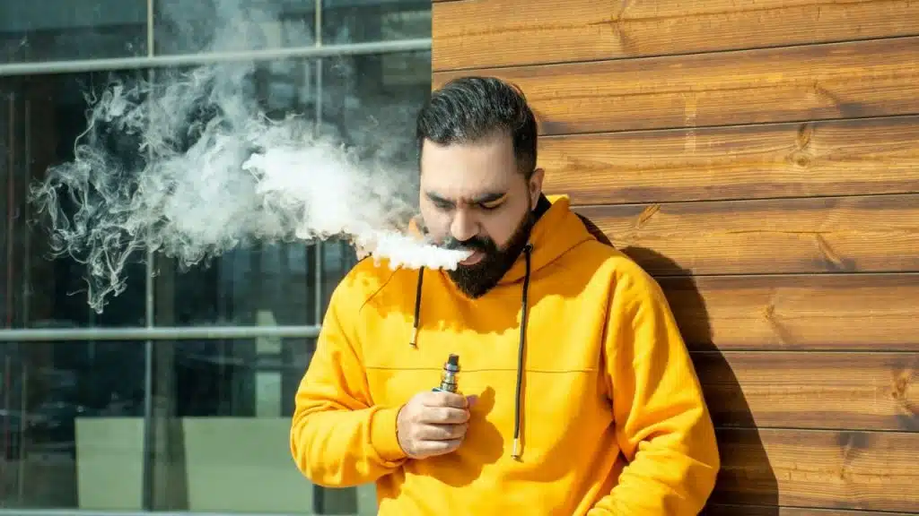 A man with a beard and mustache in a yellow hoodie smokes an e-cigarette and blows out a large cloud of vapor.
