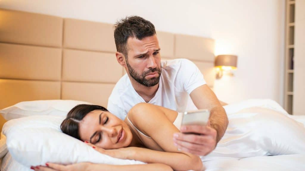 A man looks suspiciously at his partner's phone while she sleeps.
