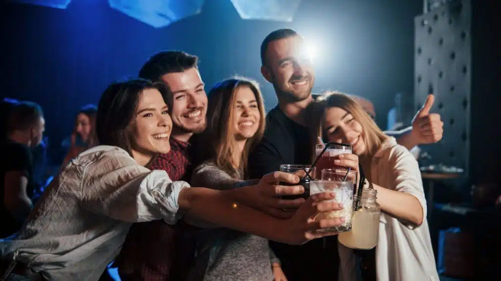 A diverse group of five friends happily toast with drinks in a dark club.