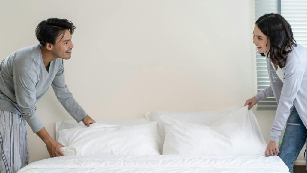 A happy couple smiles at each other while making a bed. They are both holding up a white blanket.