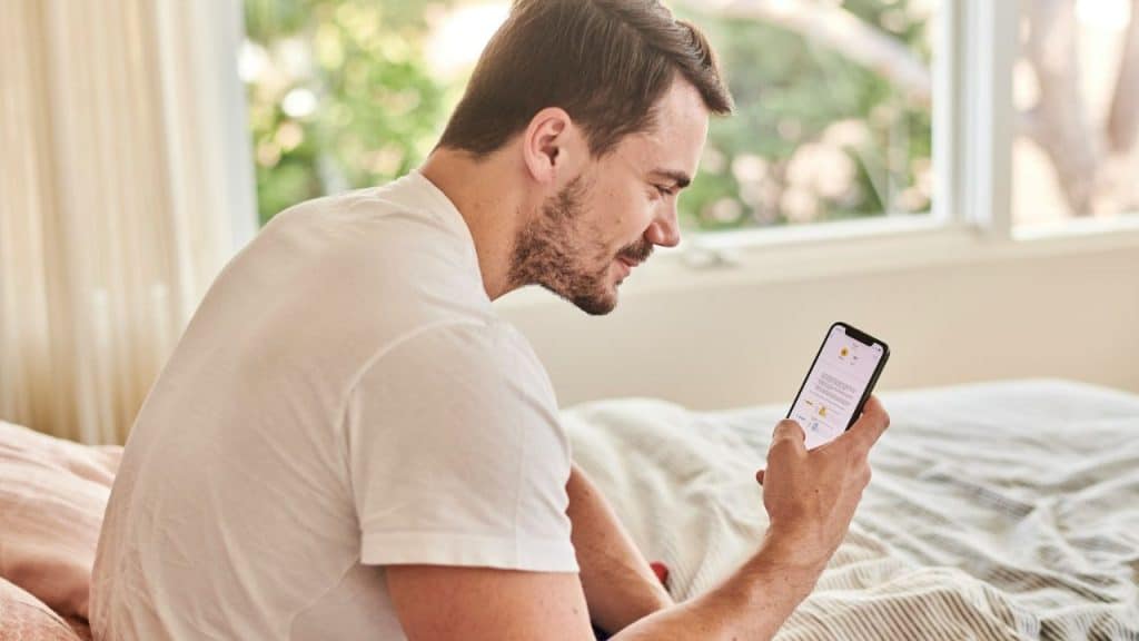 A man sits on a bed, looking at his phone and smiling.