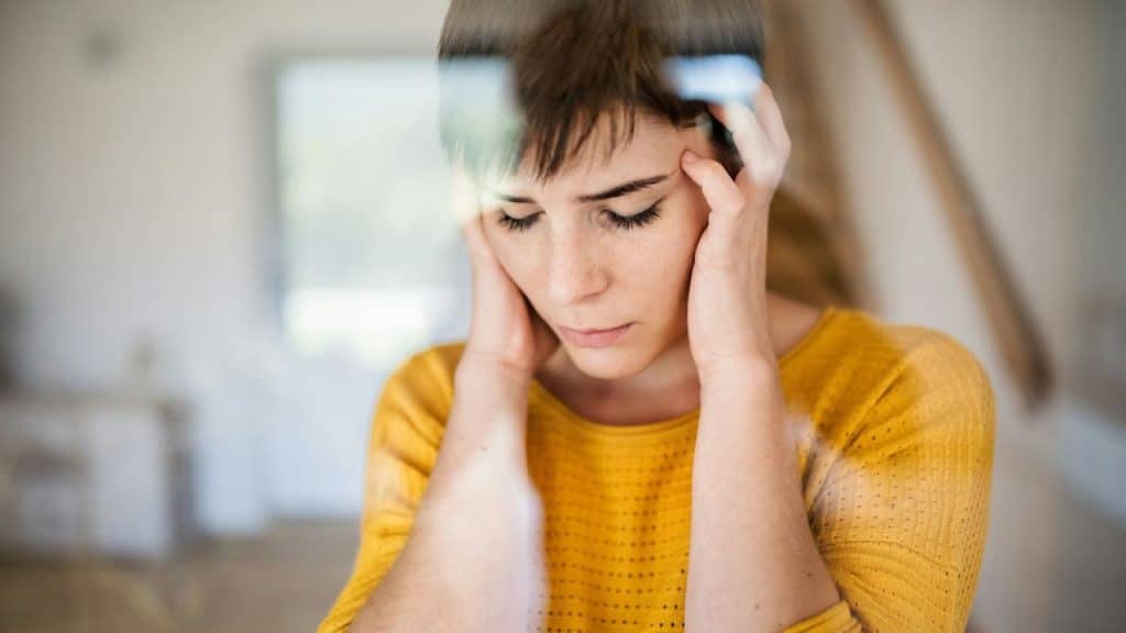 A woman in a yellow shirt with her hands to her head looks down with her eyes closed.