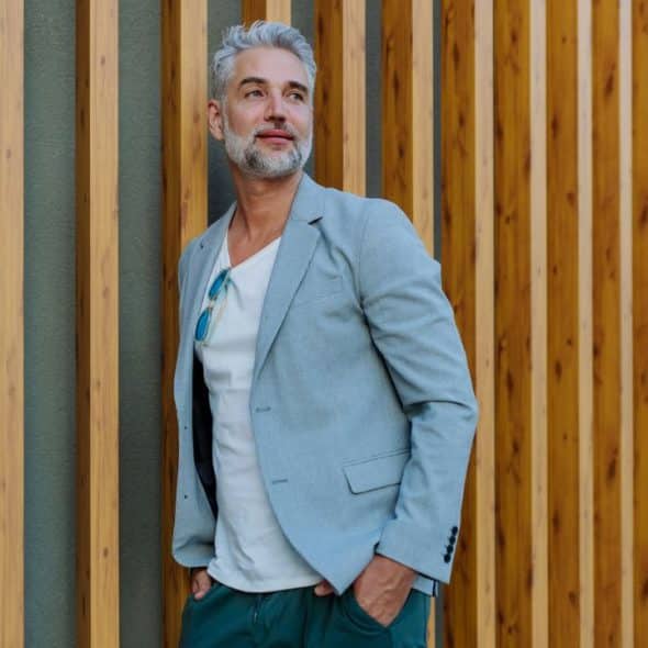 A handsome, gray-haired man in a blazer stands next to a wooden wall.