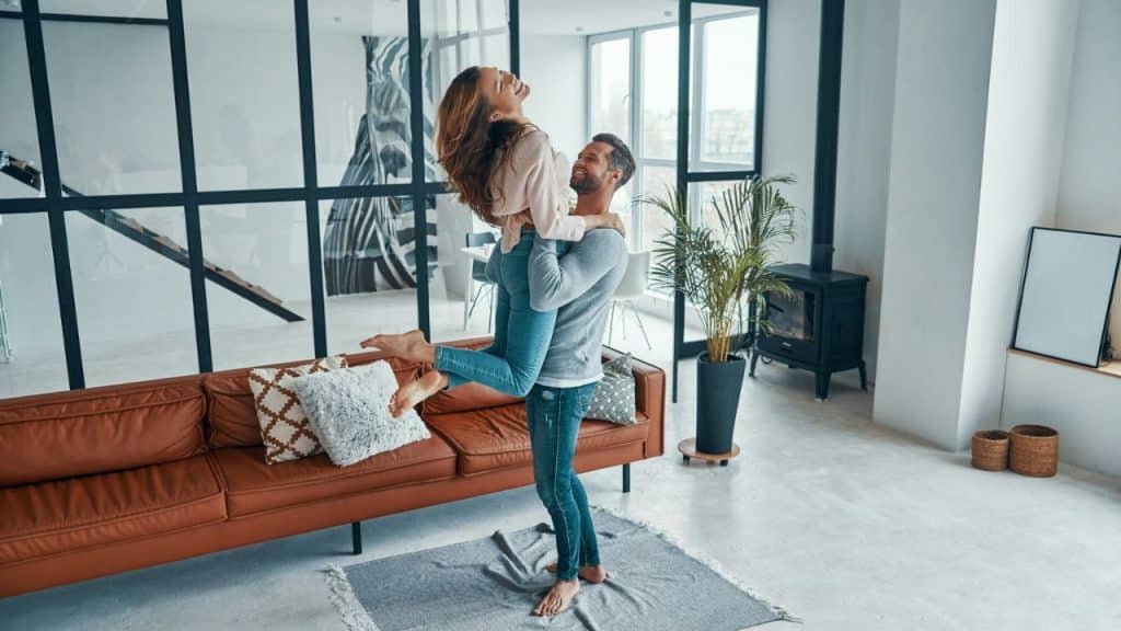 Man holding and lifting a laughing woman in a living room with a brown couch and large windows.