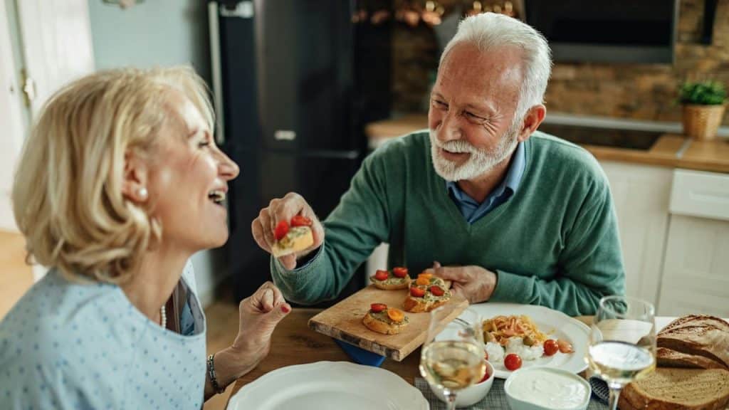 A joyful elderly couple eating bruschetta together at a kitchen table.