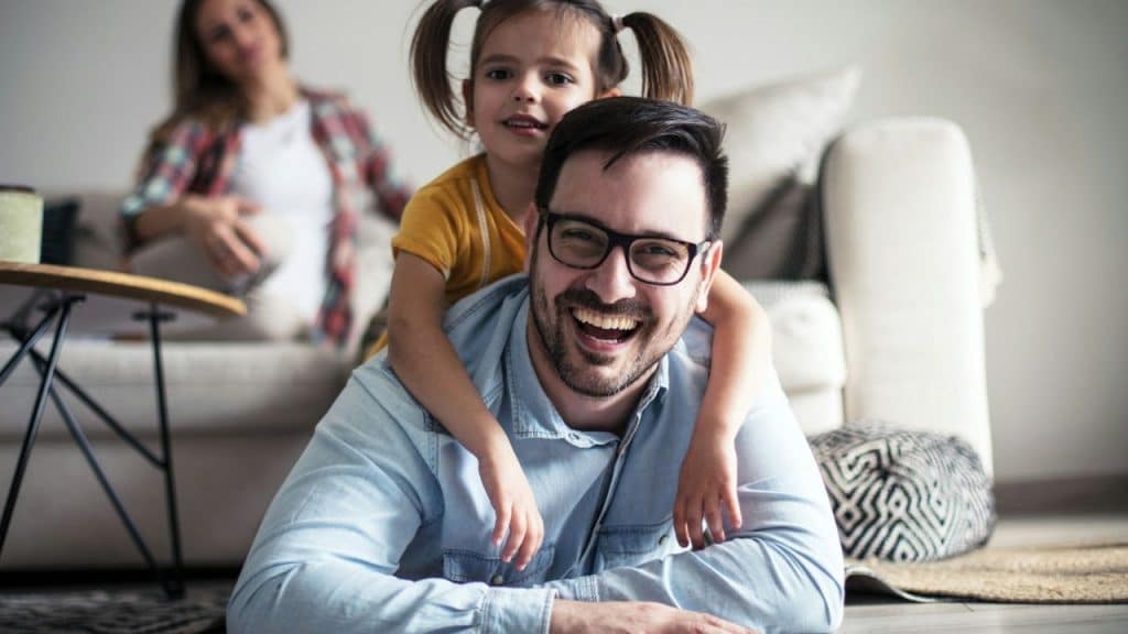 A laughing man wearing glasses is on the floor with a girl on his back indoors.