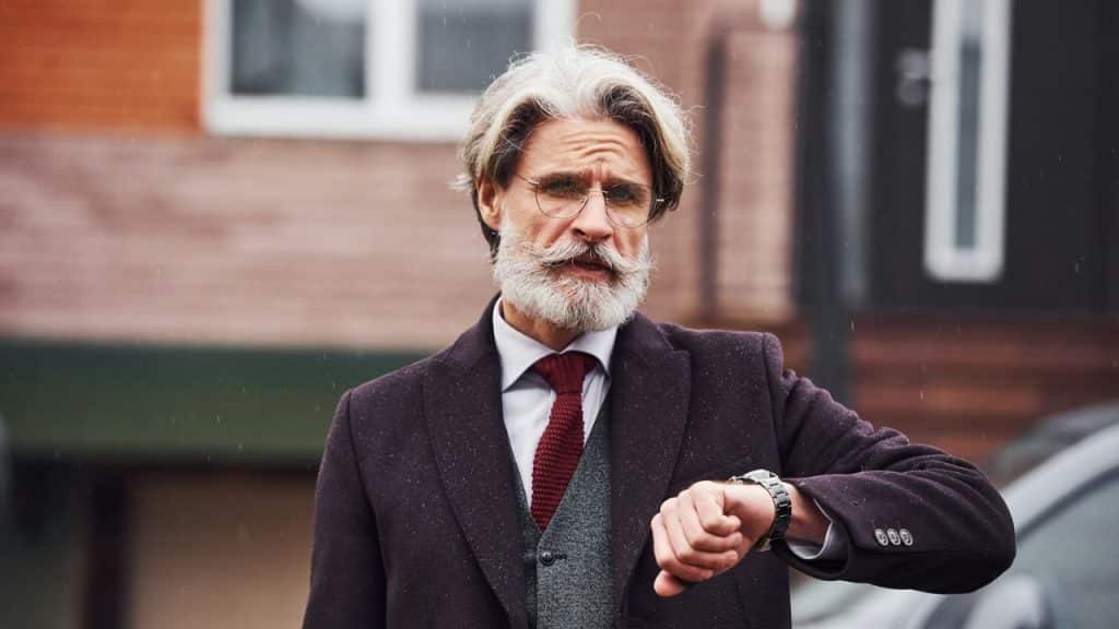 A well-groomed, older man with a gray beard and glasses looks at his wristwatch outdoors.